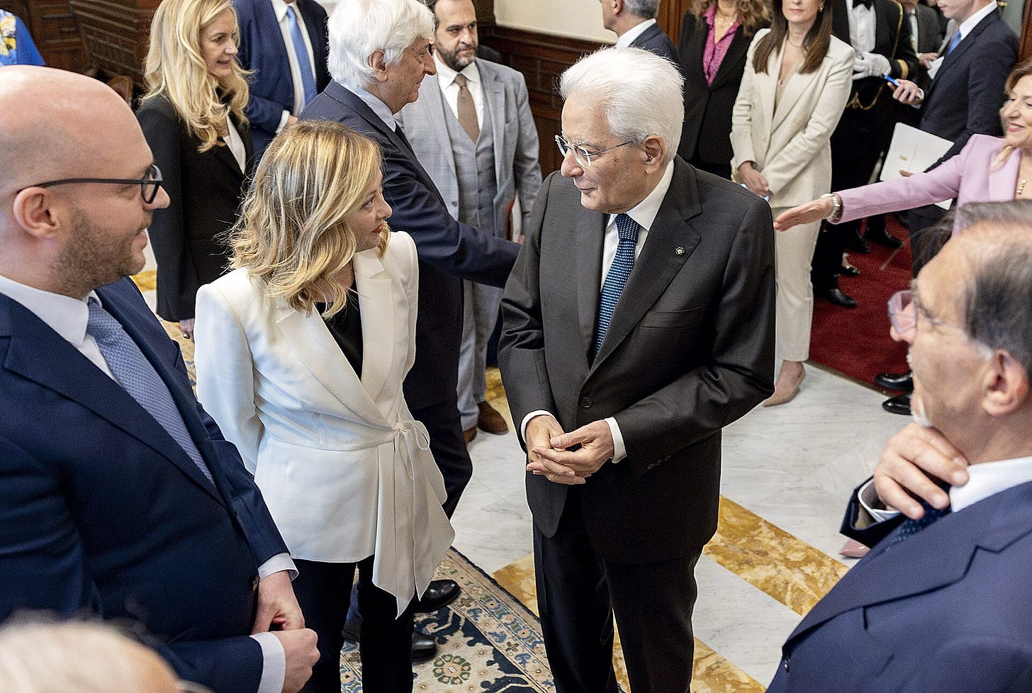 Italian President Sergio Mattarella, an elderly man in a dark suit, talks with Prime Minister Giorgia Meloni, in a white jacket, among officials in an ornate hall.