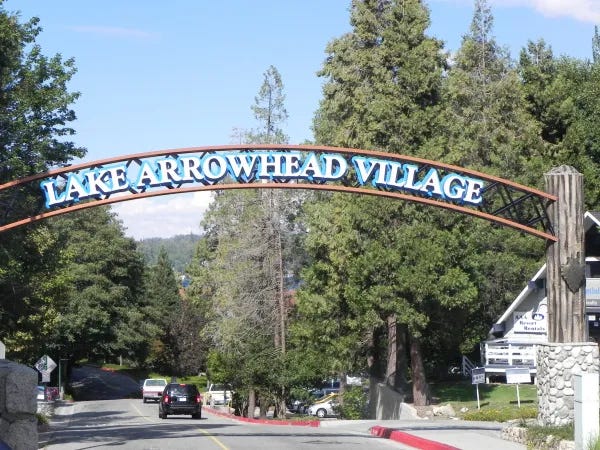 Entrance to Lake Arrowhead Village marked by a large arched sign reading “Lake Arrowhead Village” in blue letters. The sign spans a two-lane road lined with tall pine trees, with cars driving beneath it and forested hills visible in the background.