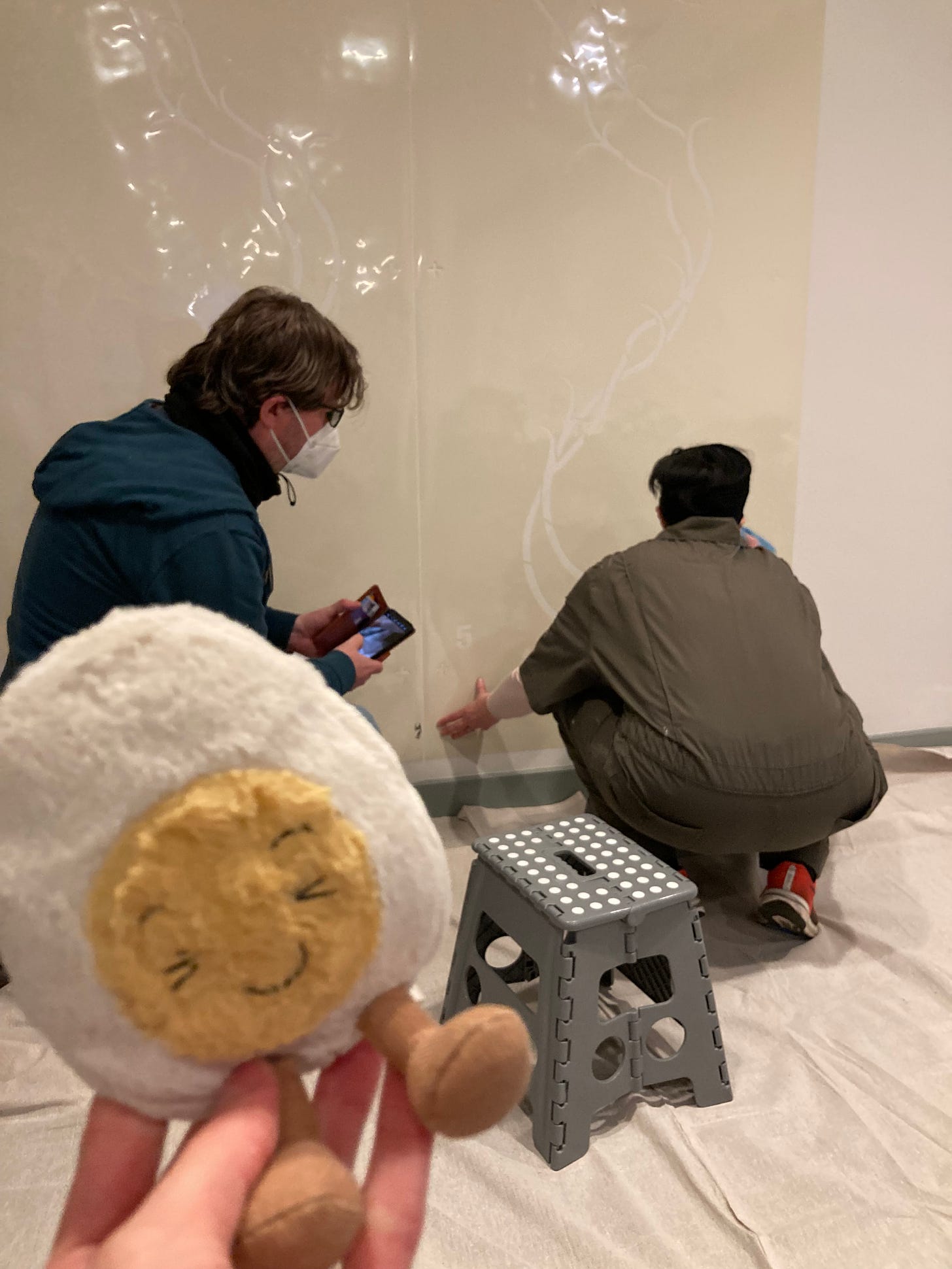 Dippy the soft toy egg in front of two people crouched on the floor wearing masks, taping up a giant stencil on the wall. The stencil cut outs are long vines running up and down the wall. 