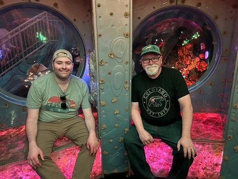 First picture shows a jumble of colorful items agains a green wall. One of the items is a trophy labeled Sports Ball. 2nd picture a young man and a man with a white beard sitting in front of two round windows. There is a green steel girder separating the sitters. #rd pic: The same two people are sitting on a couch in front of shelves with books piled haphazardly.