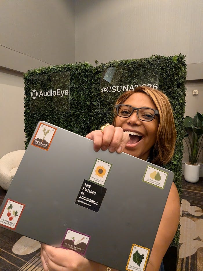 Mariella is a smiling woman with glasses stands indoors in front of a green hedge-style backdrop at a conference booth. The backdrop includes the AudioEye logo on the left and the text #CSUNATC26 on the right. She is holding up a large gray board covered with small colorful stickers. The stickers have plant or produce illustrations and accessibility-themed labels, including one black sticker in the center that reads “THE FUTURE IS ACCESSIBLE” with @projecthearing underneath. The overall image feels upbeat, playful, and celebratory, like a candid conference photo.