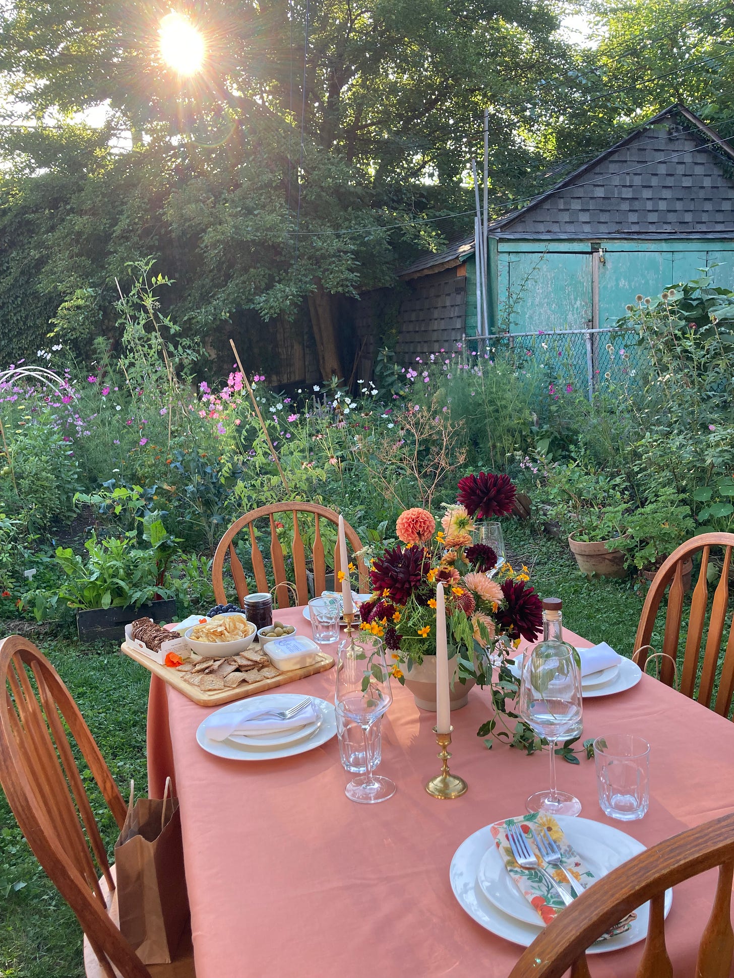 On a lawn surrounded by garden sits a fully set table with a coral coloured table cloth. On it, the table is set for formal dining, with a large floral centerpiece and a cheeseboard off to one side.