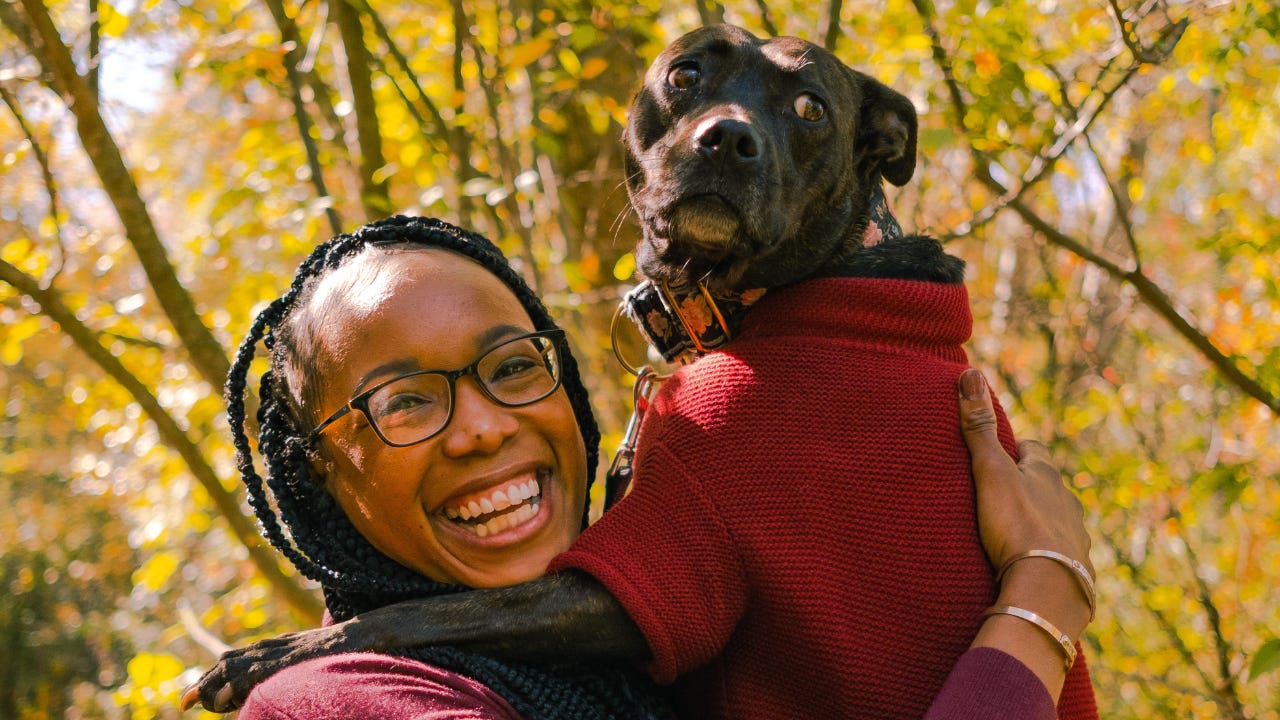 A smiling Black woman wearing glasses holds a medium-sized dog dressed in a red sweater outdoors, surrounded by autumn trees with golden leaves.