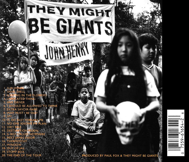Black-and-white photo of children standing in a park under a handmade banner reading “They Might Be Giants – John Henry.” One child sits in a Radio Flyer wagon while another holds a skull, creating a haunting yet playful album-back image.