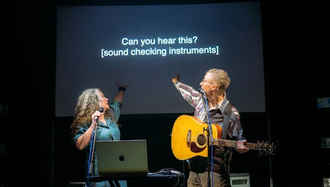 Two white people on a stage are pointing to a project screen that says Can you hear this? Sound checking instruments.