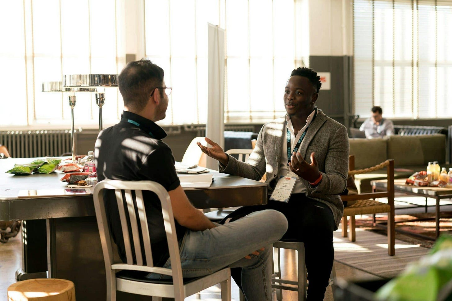 Two guys sitting on chairs in an open plan office, talking next to a large table with snacks on it.