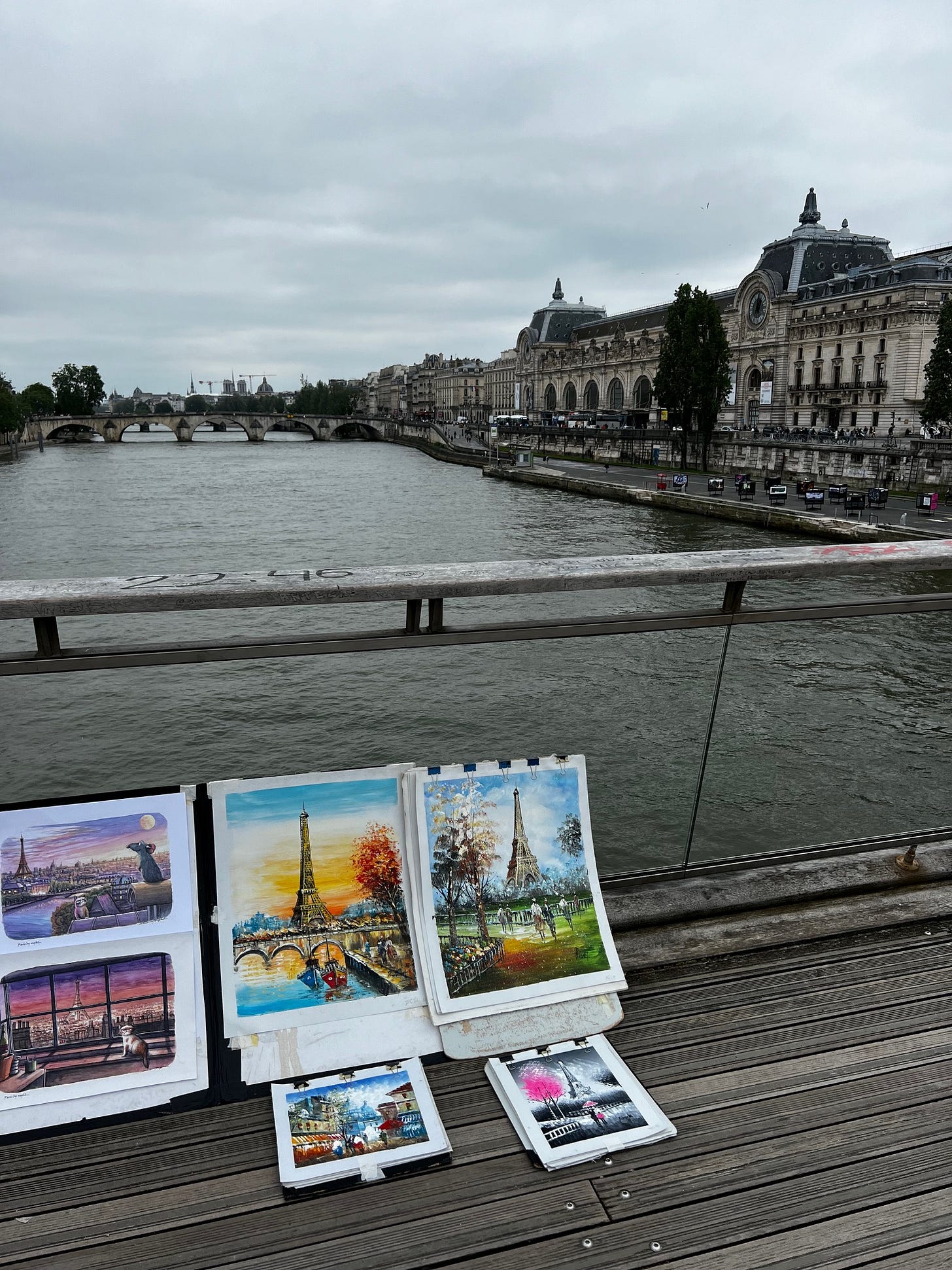 Artist's paintings displayed for sale on a wooden bridge overlooking the Seine River in Paris, with the Musée d'Orsay visible in the background. The artwork shows various Paris scenes including the Eiffel Tower and cityscapes in different styles and seasons. Artist's paintings displayed for sale on a wooden bridge overlooking the Seine River in Paris, with the Musée d'Orsay visible in the background. The artwork shows various Paris scenes including the Eiffel Tower and cityscapes in different styles and seasons.