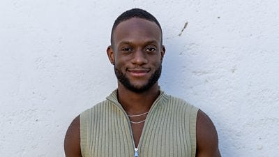 Aron wears a knitted zip vest in a sage green colour and two silver chains