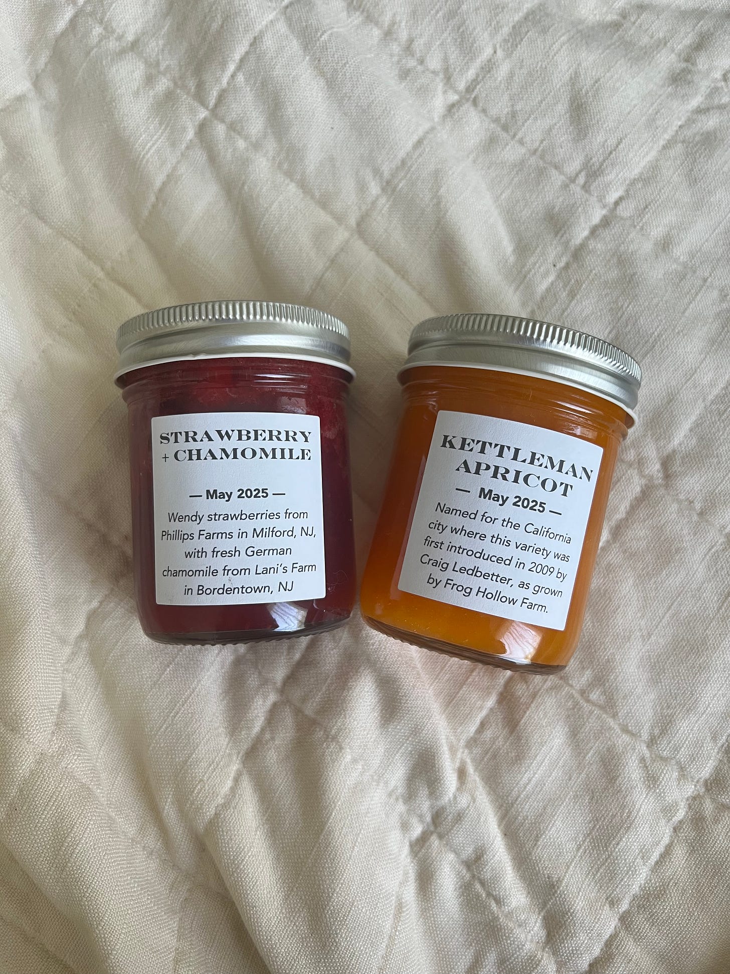 there are 2 glass jars full of jam, one is a bright red strawberry chamomile and the other is a peachy orange kettleman apricot. they are laid out on a quilt. 