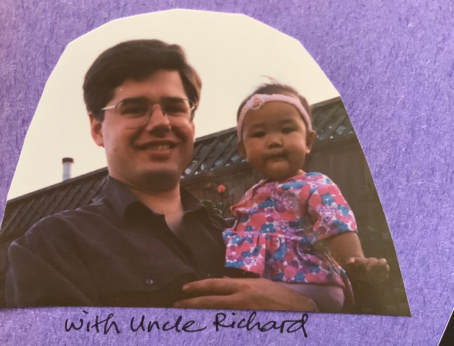Picture of a piece of a scrapbook family photo album, on purple construction paper, showing Uncle Richard holding baby me. He's wearing rectangular glasses and a dark button up shirt, and I'm wearing a floral dress and a pink satin baby headband with a small rosette on it. He's smiling and I've got a baby expression that's a little confused. Circa May 1992.