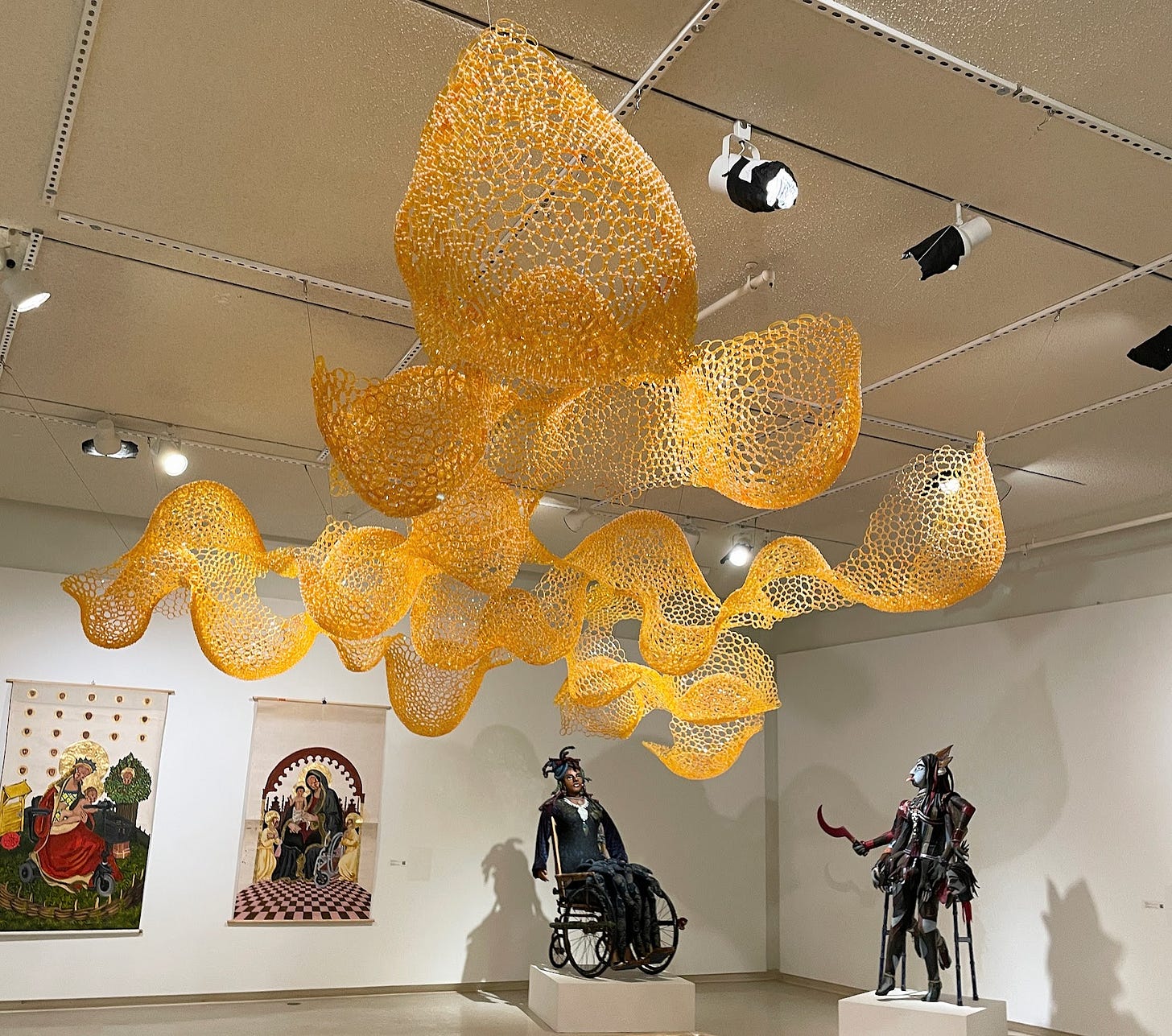 Gallery with an undulating golden ceiling installation of prescription bottle slices cable tied together into bubbles and swirls. Below are two wheelchair madonnas, a sculture of Medusa built into a vintage wheelchair, and a multi-limbed, multicolored goddess with three eyes who is built into a walker.