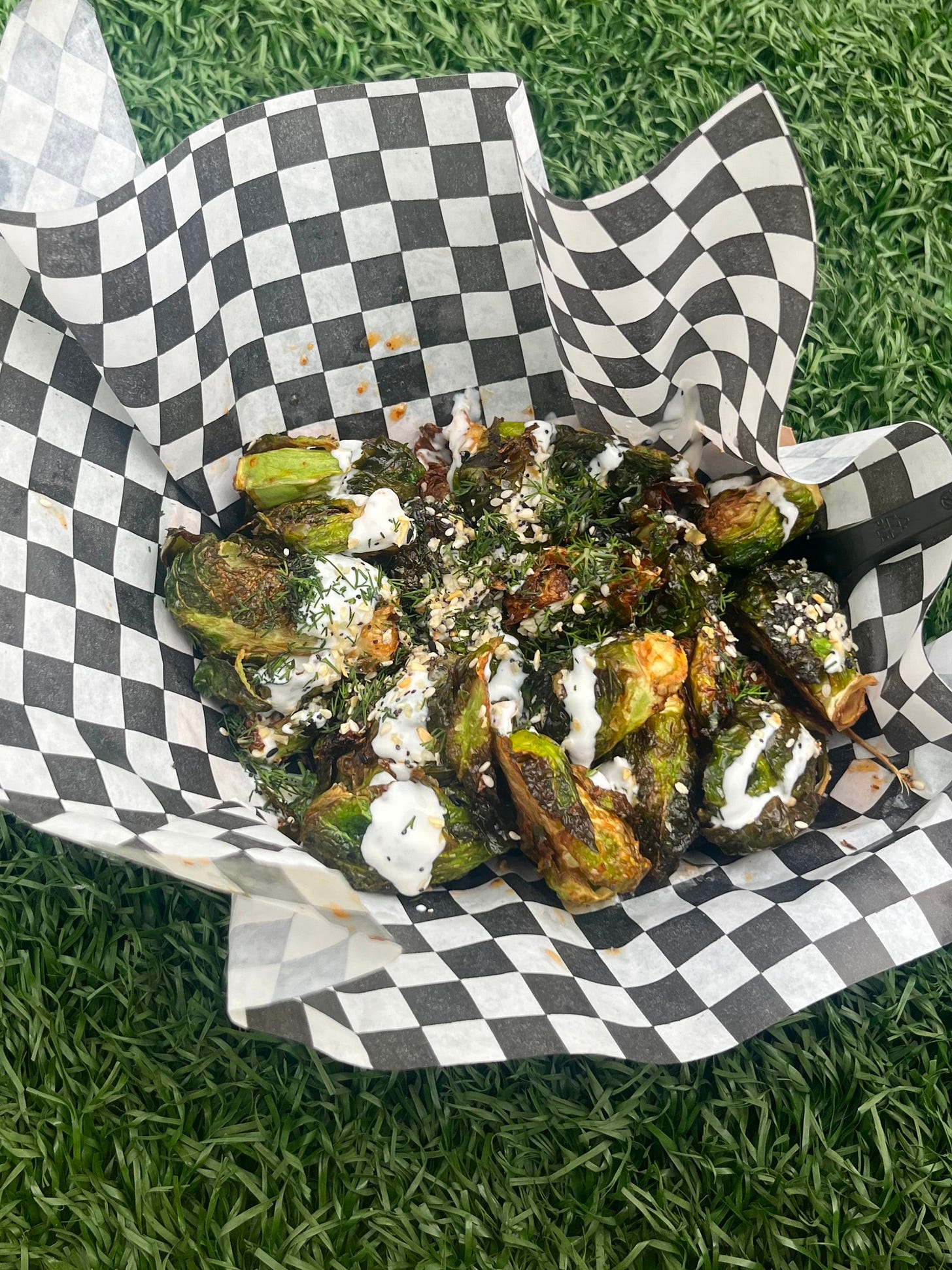 Roasted brussels sprouts covered with an aioli, fresh dill, and everything bagel seasoning, wrapped in black and white checkerboard paper and resting on astroturf.
