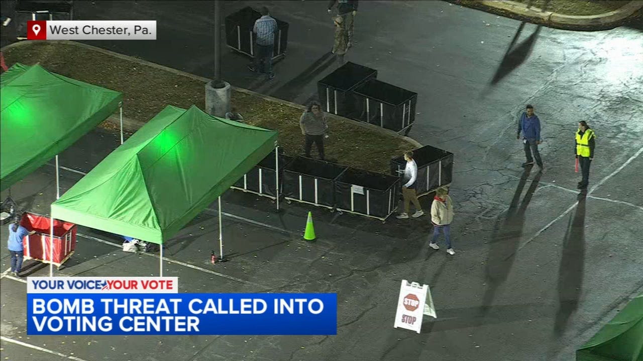 A picture of a parking lot in West Chester, Pennsylvania. It is a screenshot from a news report. The chyron reads Bomb Threat Called Into Voting Center. There are election workers on the ground hauling around ballots and green tents.