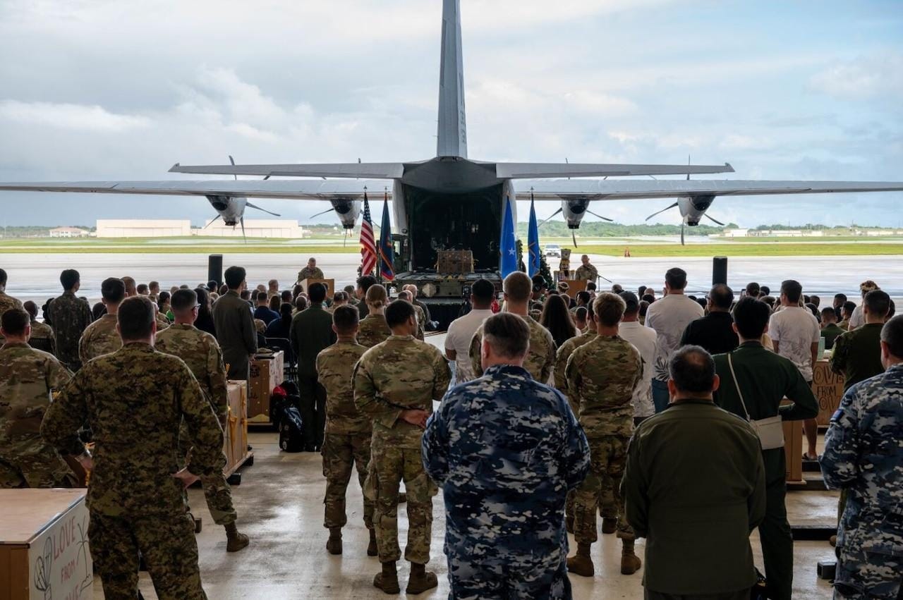 A group of service members and civilians stand behind an open military aircraft in a hangar.