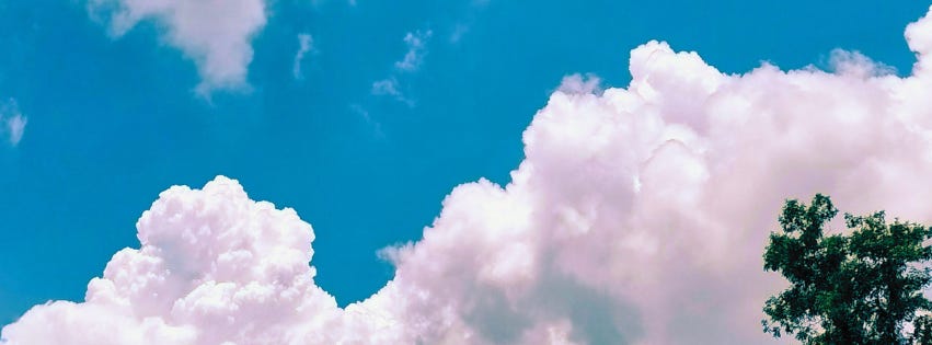 Vibrant blue sky with large fluffy white clouds and a glimpse of green tree foliage in the corner.