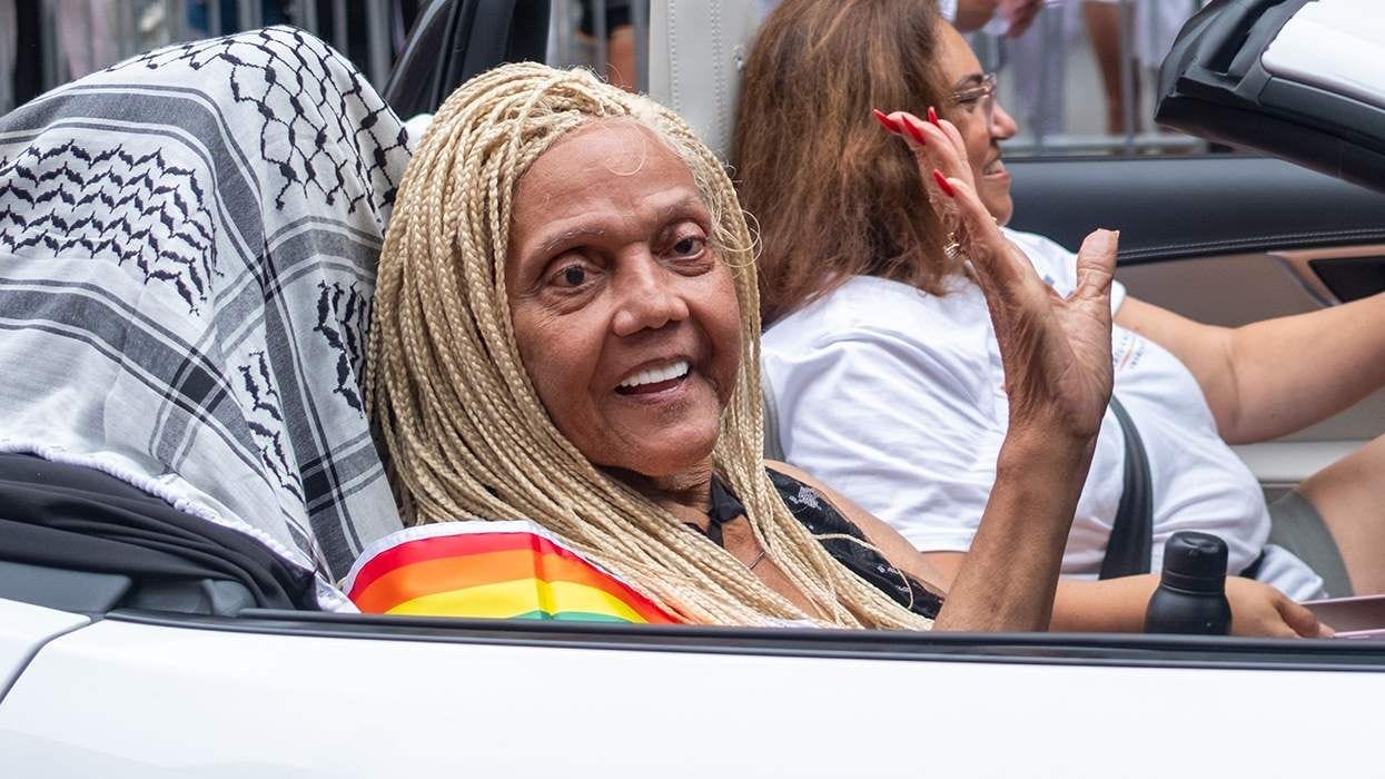 Miss Major with a rainbow top, blonde braids, and bright red acrylic nails waving from a convertible as the Grand Marshall of NYC Pride festivities 2024, riding with a Palestinian kuffiyeh draped over the seat behind her