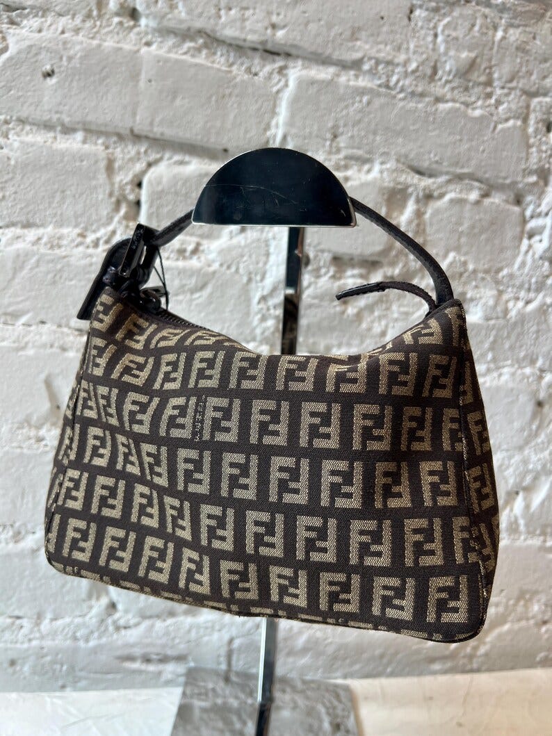 May include: A small brown and beige handbag with a brown leather strap. The bag is made of a fabric with a repeating pattern of the letter 'F'. The bag is hanging on a metal stand.