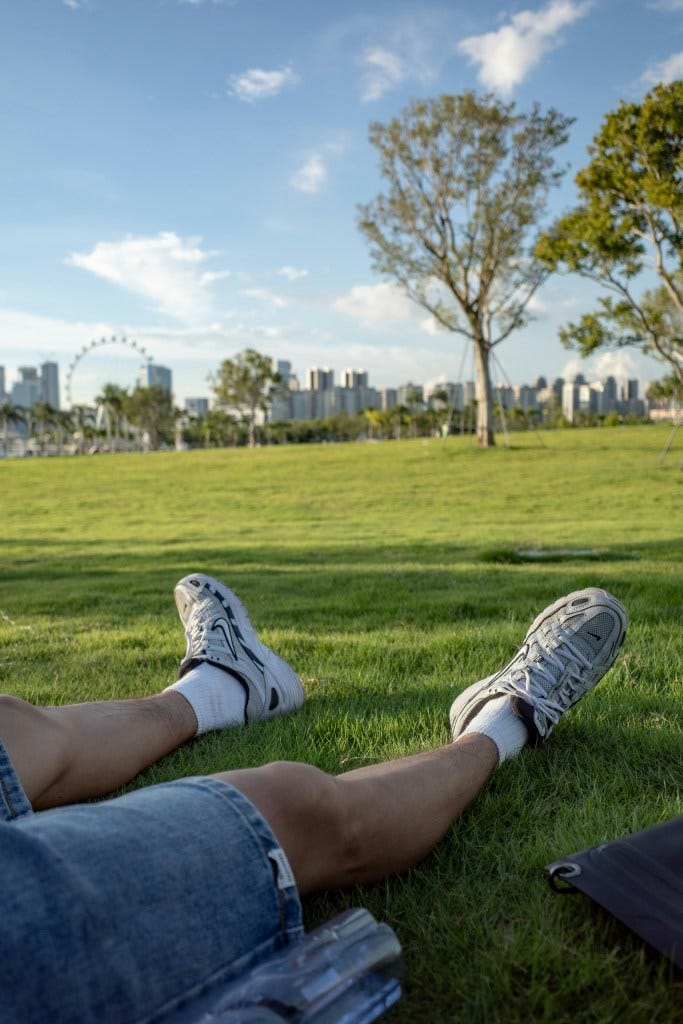 A person relaxing on grass, with their legs stretched out wearing denim shorts and sneakers, overlooking a park with trees and a city skyline in the background.