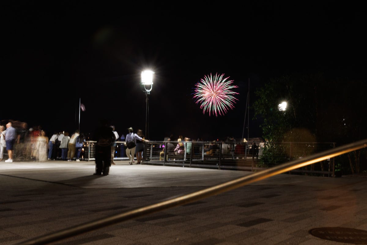 Large green and red firework above Boston Harbor with motion-blurred crowd on pier watching