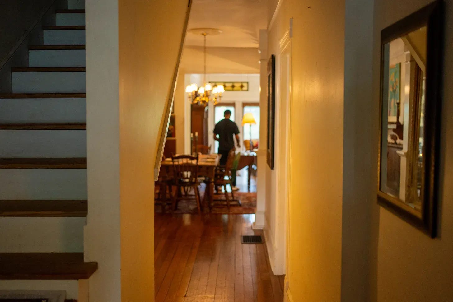 View down a hallway with hardwood floors looking into a softly lit dining room where a person is standing by a set table.