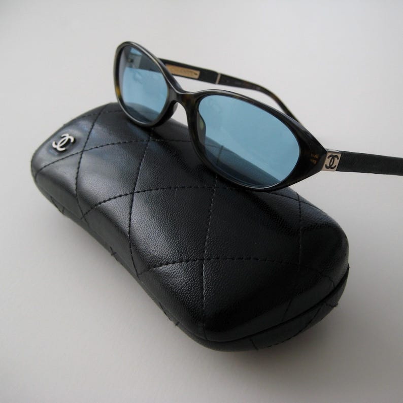 May include: Black sunglasses with blue lenses and a black leather case with a quilted pattern and a silver Chanel logo.