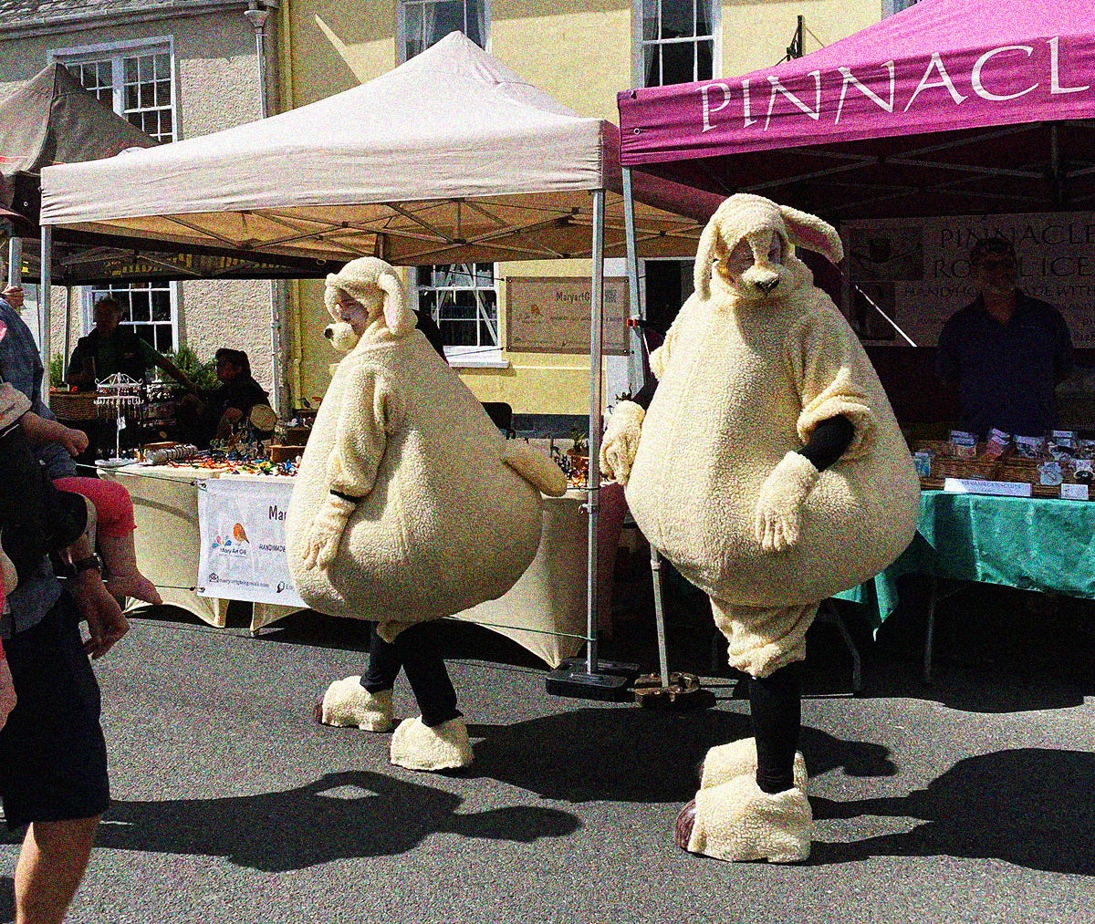 Two grown adults dressed as cartoon sheep  looking at market stalls in the sunshine