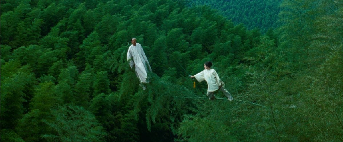 Two characters in white, holding weapons and confronting one another, standing aloft amid the treetops of a forest. Still from the film "Crouching Tiger, Hidden Dragon."