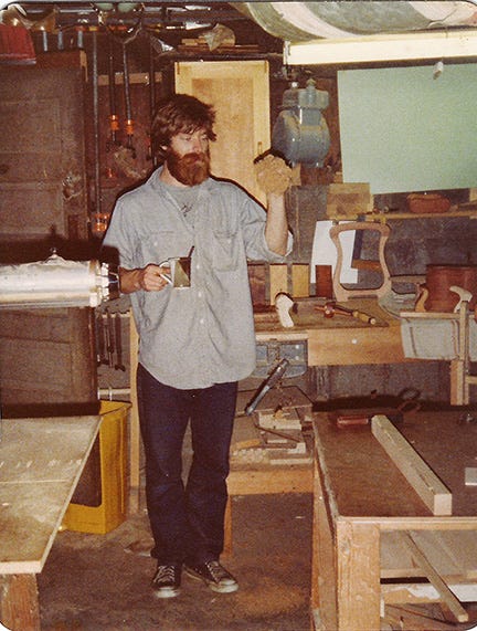 Young Gary Rogowski standing in his first woodworking shop in 1977, holding a tool and a piece of wood amid benches and tools.