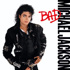 A man in his late 20s stands and looks forward. His hair is curly and black. He is wearing a black jacket that has several buckles and pants. The background is white and beside him are the words "Michael Jackson" in black capital letters, and over them, "Bad" in red.