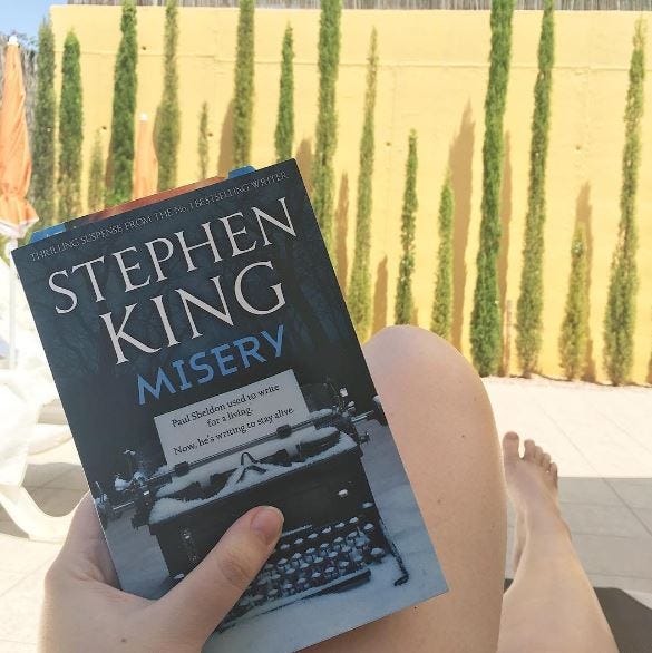 A girl on a sun lounger holding a paperback copy of Misery by Stephen King. The book cover is of a typewriter covered in snow on a blue background. A girl on a sun lounger holding a paperback copy of Misery by Stephen King. The book cover is of a typewriter covered in snow on a blue background.