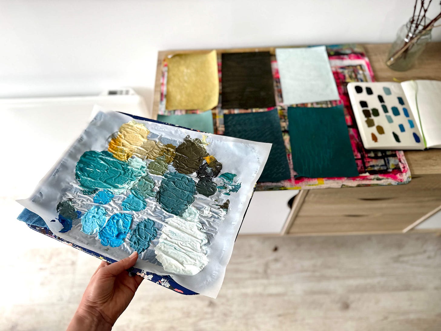 Hand holding a wet Palette with acrylic paint and collage papers and sketchbook arranged on a sideboard