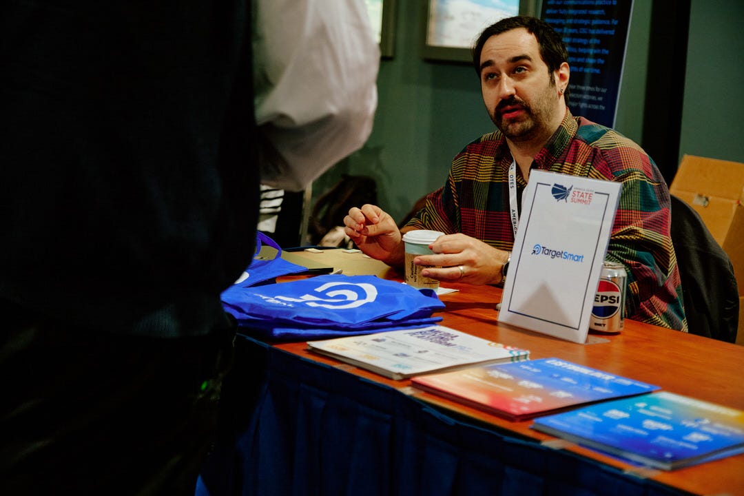 Man in multicolored plaid flannel shirt behind a vendor table with TargetSmart signage and blue branded tote bags in the foreground, mid-conversation with someone off frame with a coffee cup in hand