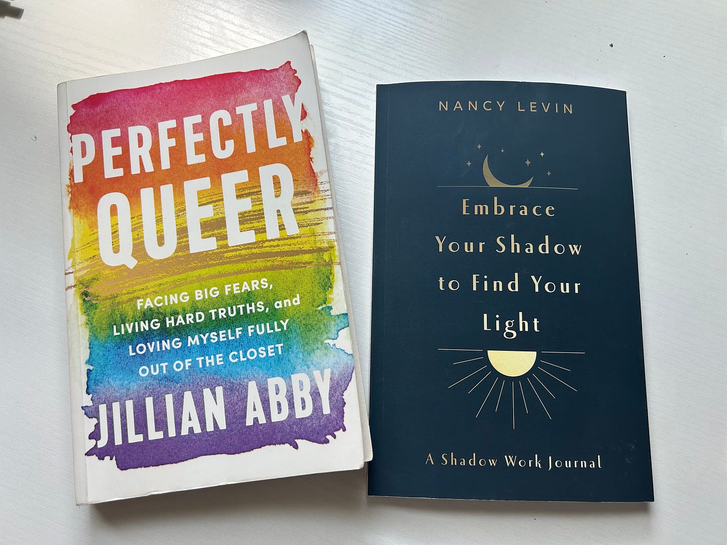 Two books, one of Perfectly Queer by Jillian Abby with a rainbow cover and "Embrace Your Shadow to Find Your Light" by Nancy Levin with a navy cover. Two books, one of Perfectly Queer by Jillian Abby with a rainbow cover and "Embrace Your Shadow to Find Your Light" by Nancy Levin with a navy cover.