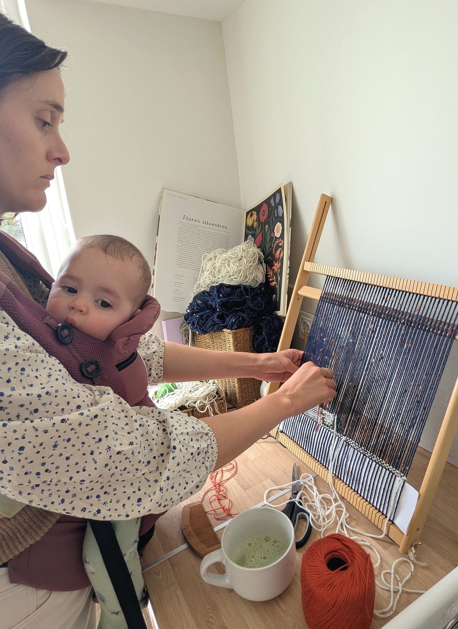 Rebecca weaving at a small loom with a baby in a carrier on her chest, working one-handed at a table with yarn and tools. Rebecca weaving at a small loom with a baby in a carrier on her chest, working one-handed at a table with yarn and tools.