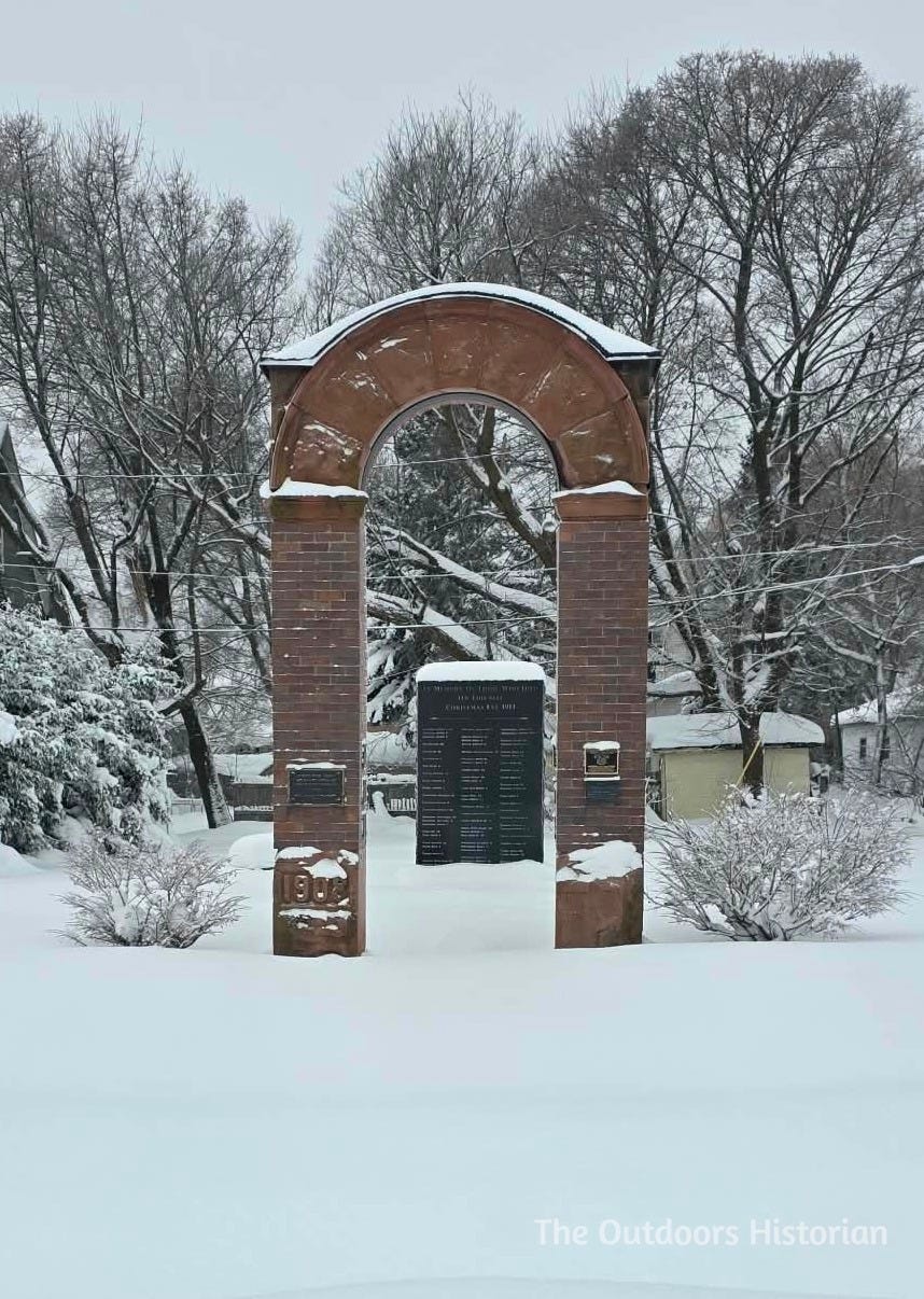 A close-up of the red sandstone archway at the Italian Hall memorial site in December. The stones are dusted with snow, and the arch stands as a lone, silent monument in the white landscape.