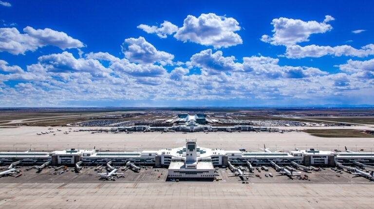 An external shot of Denver International Airport (DEN) with teh airport in the foreground, with planes parked up at the gates and the horizon in the distance and a blue sky above this