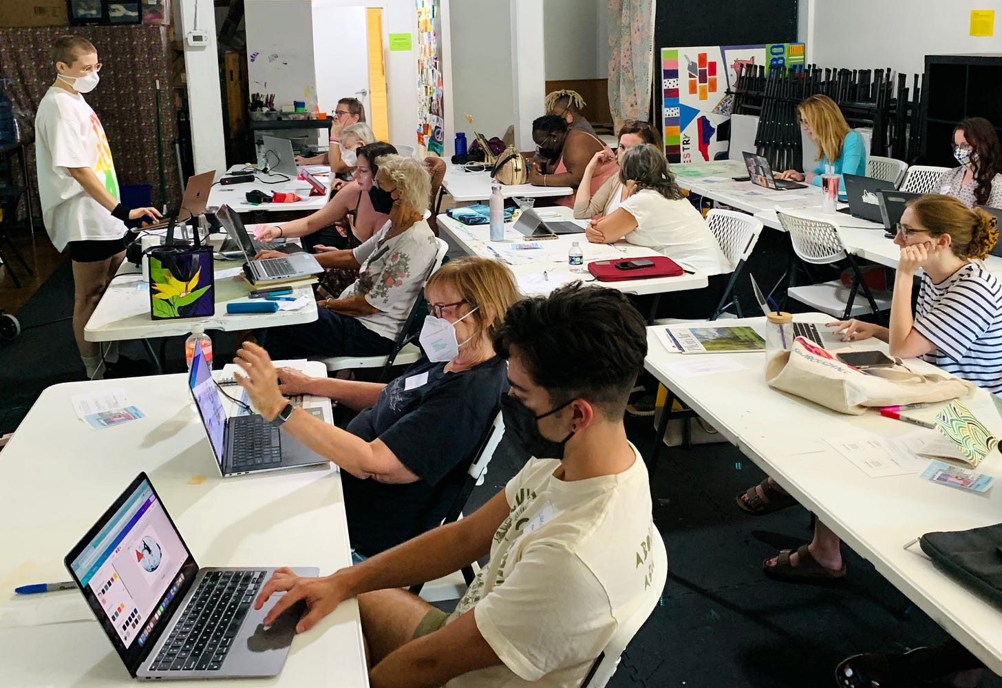 A room of people on laptops wearing masks working on Canva, Shady teaching at the front of the room.