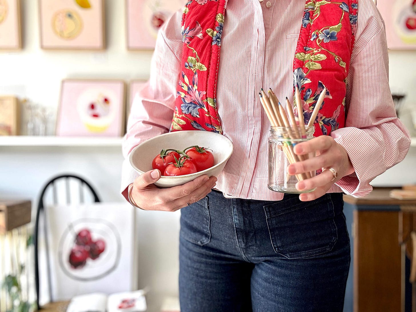 A close-up shot of an artist in a bright studio, holding a small ceramic bowl filled with fresh vine tomatoes in one hand and a glass jar of coloured pencils in the other. The artist wears a pink striped shirt, dark denim trousers, and a vibrant red floral quilted vest. In the soft-focus background, framed mixed-media artwork on pink-primed pages is displayed on a gallery wall. A close-up shot of an artist in a bright studio, holding a small ceramic bowl filled with fresh vine tomatoes in one hand and a glass jar of coloured pencils in the other. The artist wears a pink striped shirt, dark denim trousers, and a vibrant red floral quilted vest. In the soft-focus background, framed mixed-media artwork on pink-primed pages is displayed on a gallery wall.