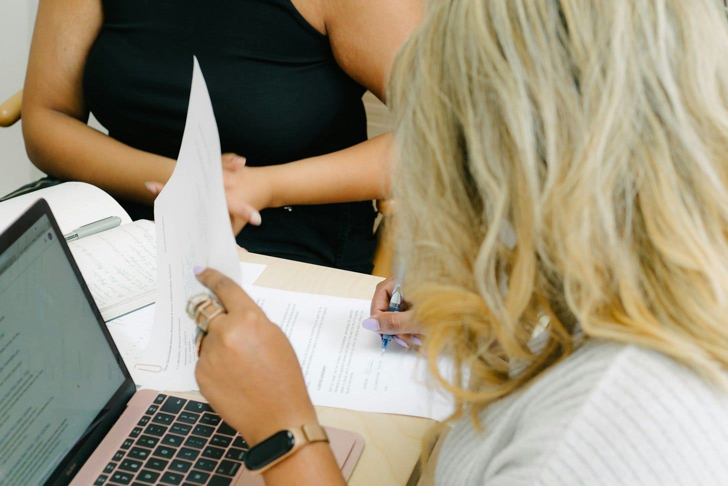 A woman reads a printed page with a pen in hand beside her open laptop, while a colleague sits across the table. A woman reads a printed page with a pen in hand beside her open laptop, while a colleague sits across the table.