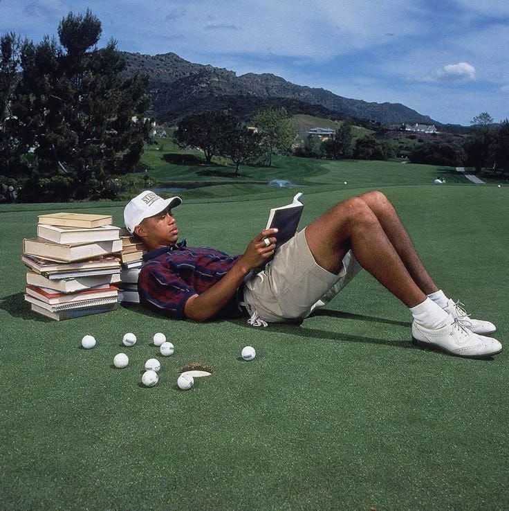This may contain: a man laying on the ground reading a book next to piles of books and golf balls This may contain: a man laying on the ground reading a book next to piles of books and golf balls