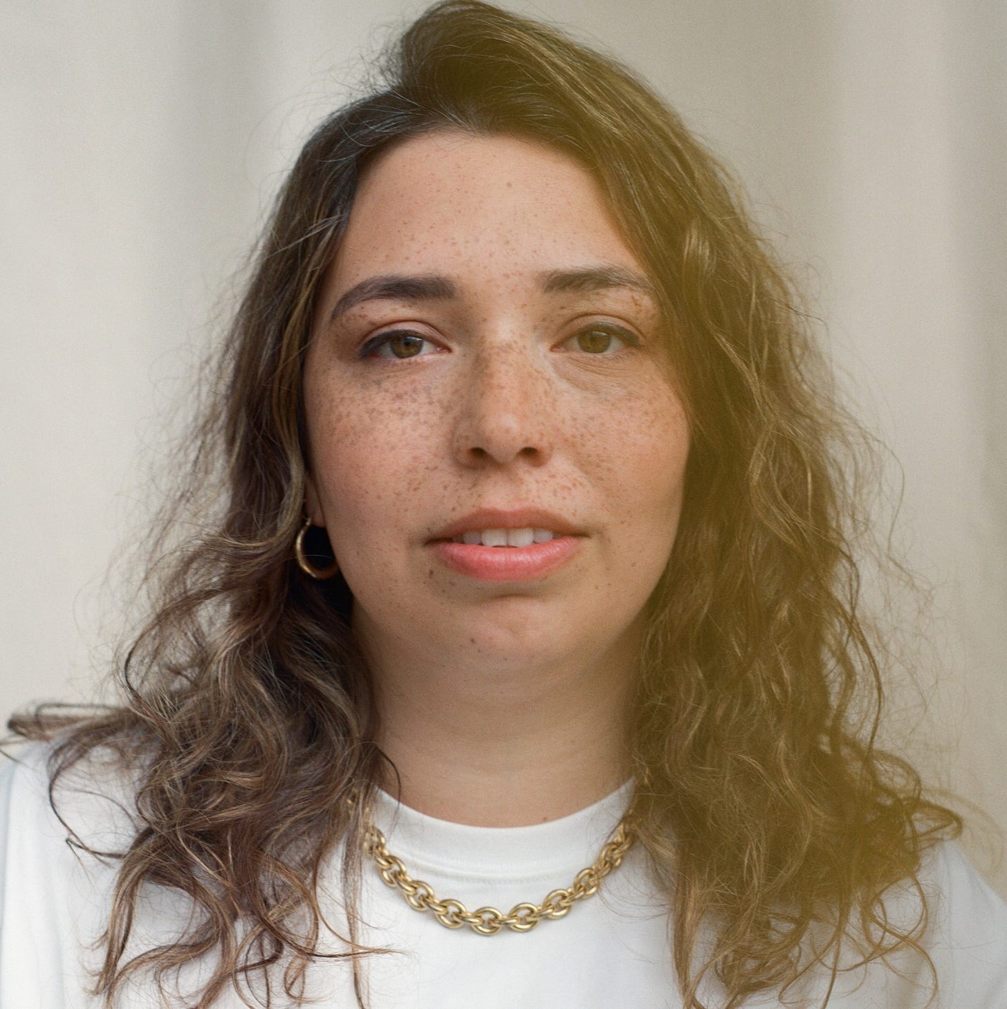 Portrait of writer Emma van Straaten, a British-Mauritian novelist, photographed by Robin Christian, showing her with wavy brown hair, light freckles, gold hoop earrings, and a gold chain necklace, wearing a white top against a soft neutral background, used for her interview with Finbarre Snarey from Tarot DMs.