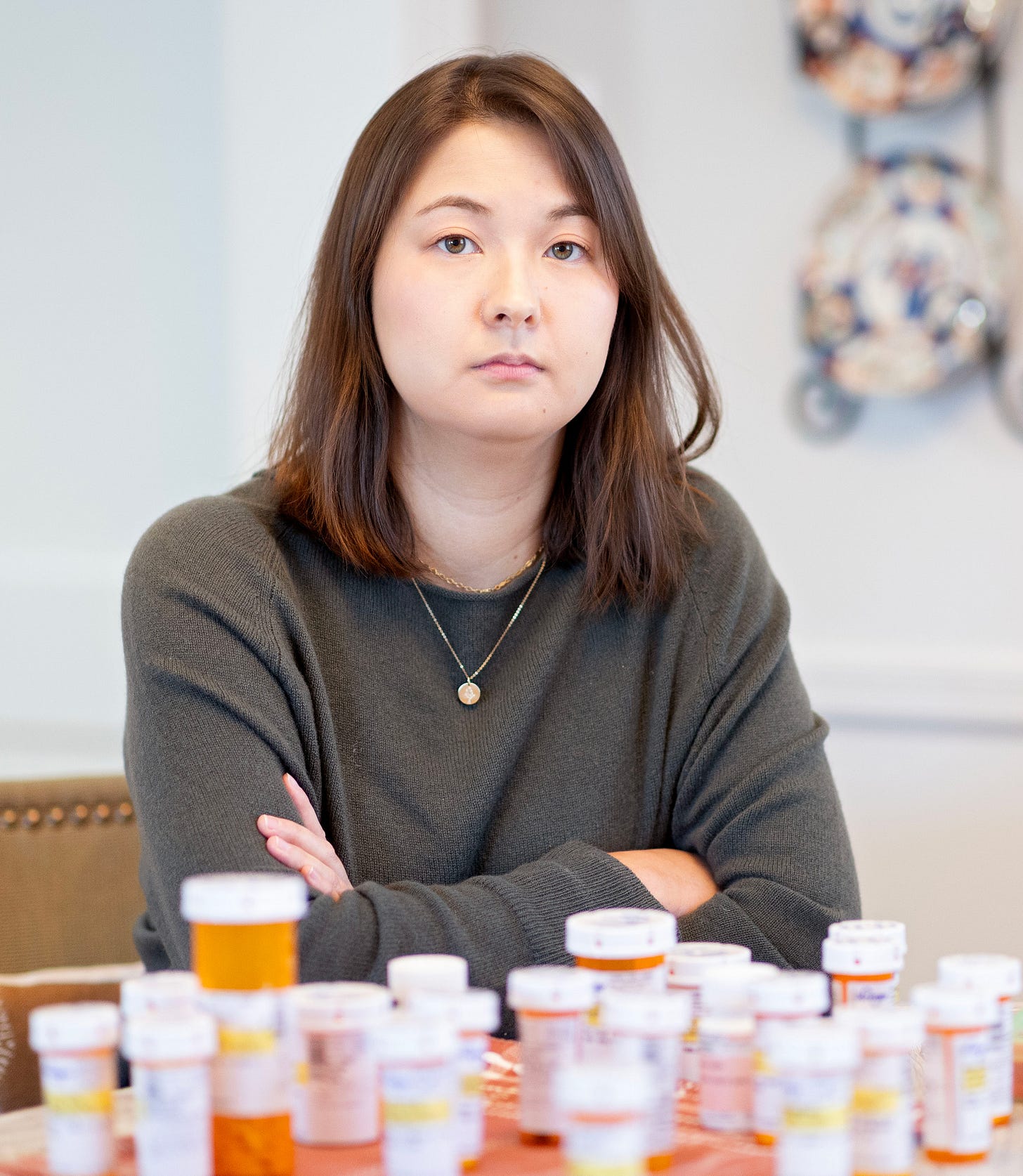 A photo of a woman sitting at a table with over a dozen pill bottles in front of her. A photo of a woman sitting at a table with over a dozen pill bottles in front of her.