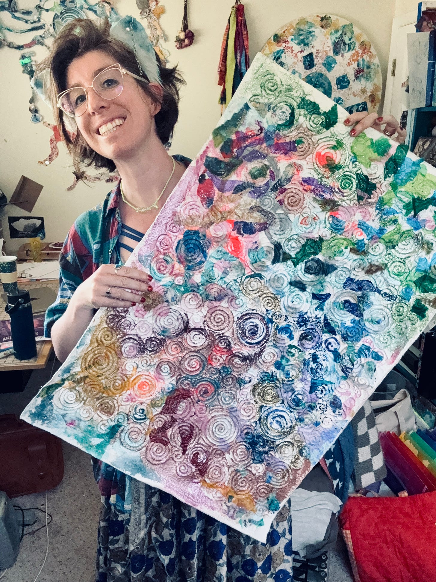 Khiri holding a piece of spiral embossed paper covered in a rainbow of art materials. She is smiling and standing in her messy studio.