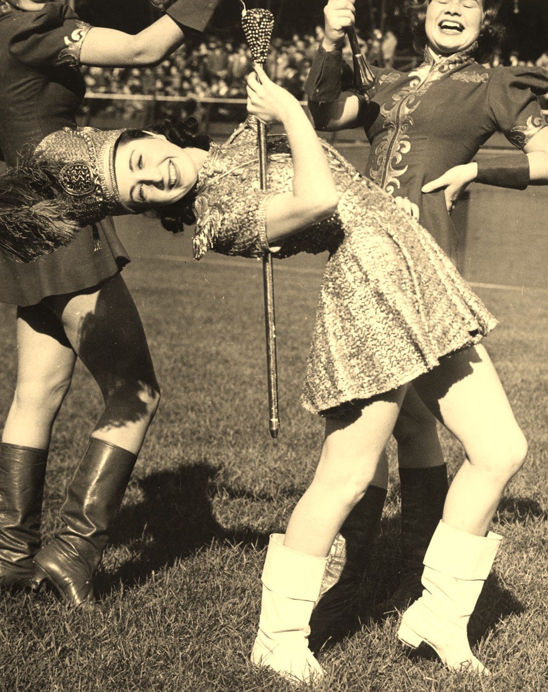 lead majorette, Rita Phillips, doing a back bend while holding a baton; two other majorettes marching on field behind Phillips lead majorette, Rita Phillips, doing a back bend while holding a baton; two other majorettes marching on field behind Phillips