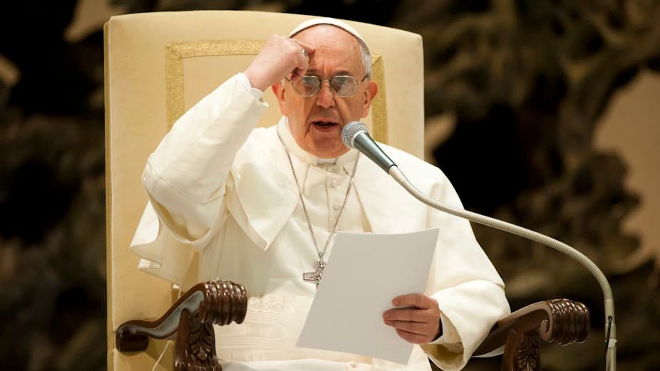 A man in a white robe speaks into a microphone while seated