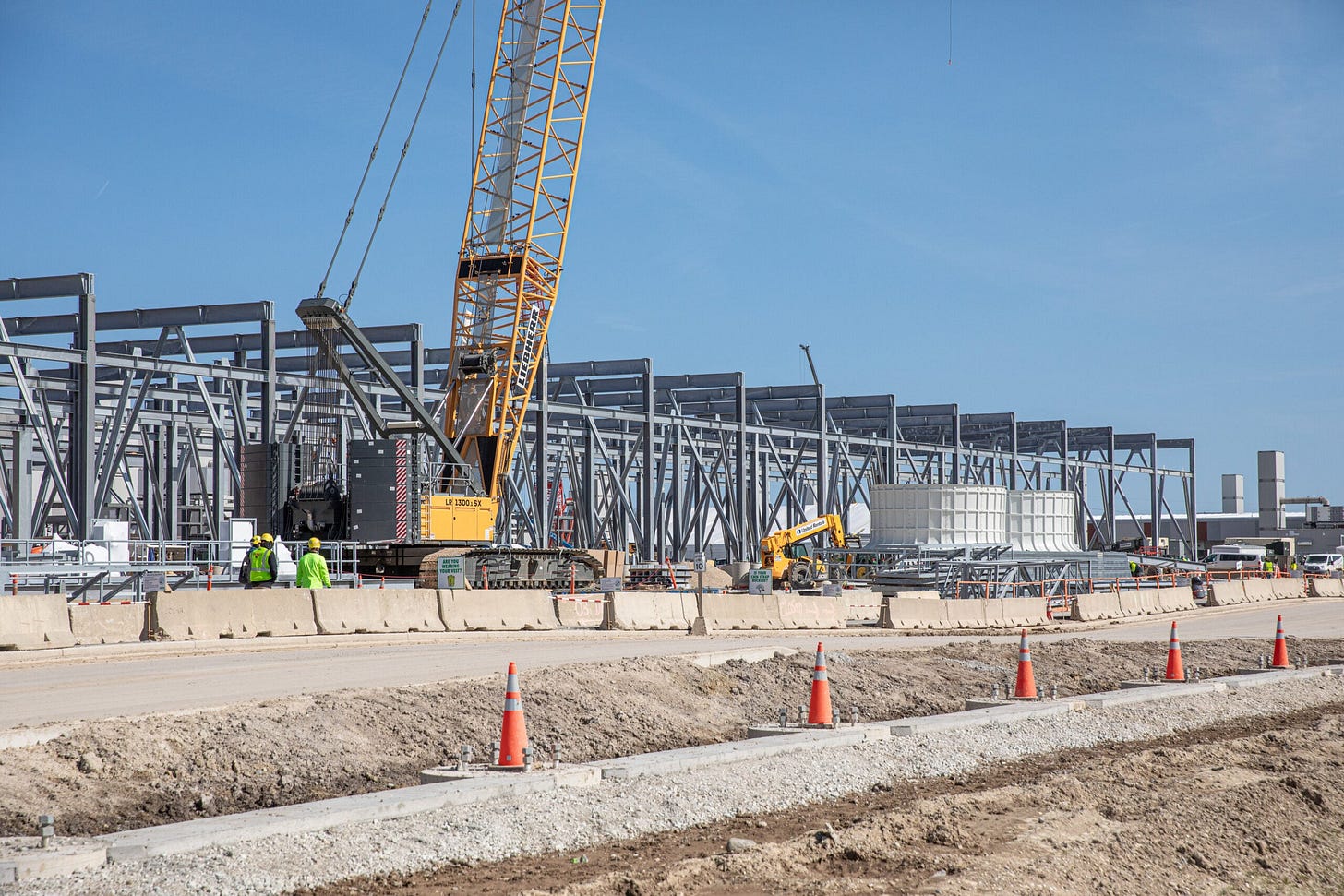 Construction site with large steel framework, a crane, and several workers in safety vests. Orange traffic cones line the foreground, and the sky is clear. Construction site with large steel framework, a crane, and several workers in safety vests. Orange traffic cones line the foreground, and the sky is clear.