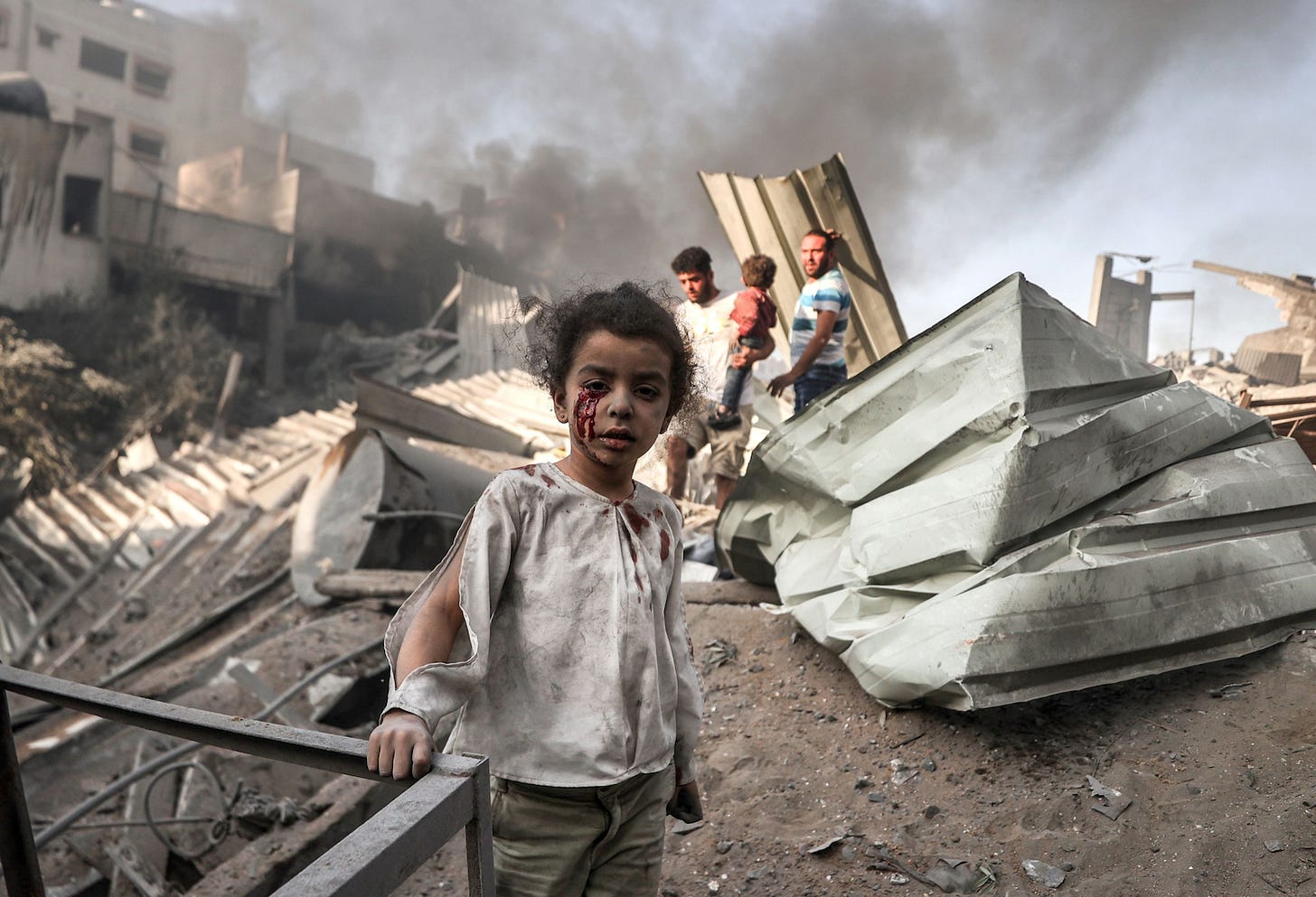 A bloodied Palestinian child stands in front of rubble and smoke after an Israeli bombardment.