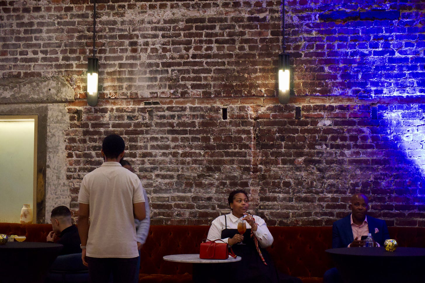 A woman sits alone at a small round marble table against a red velvet banquette in a dimly lit brick-walled venue. She holds a cocktail glass near her lips, looking slightly off to the side. A bright red handbag rests on the table in front of her. Other guests are nearby, but she appears momentarily solitary beneath warm wall sconces and blue accent lighting.