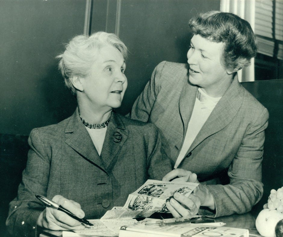 An image of two people, one seated at a desk, and the other standing, smiling and looking at each other. Both individuals are dressed in business attire.