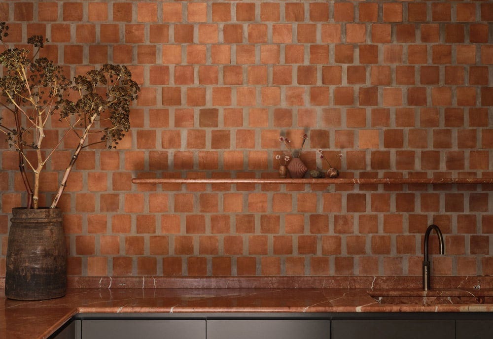 A kitchen scene featuring a textured terracotta tile wall, a wooden vase with dried branches, and a marble countertop with a sleek faucet. A kitchen scene featuring a textured terracotta tile wall, a wooden vase with dried branches, and a marble countertop with a sleek faucet.